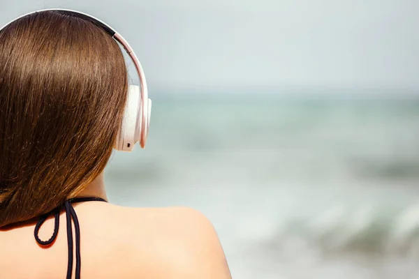 Back view of alone woman sitting on a beach in headphones listen music dreaming and looking on the sea. Female relaxation at summer vacation.