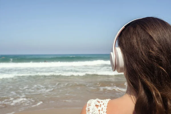 Alone woman on a beach in headphones listen music looking on the sea. Female relaxation at summer vacation. Back view