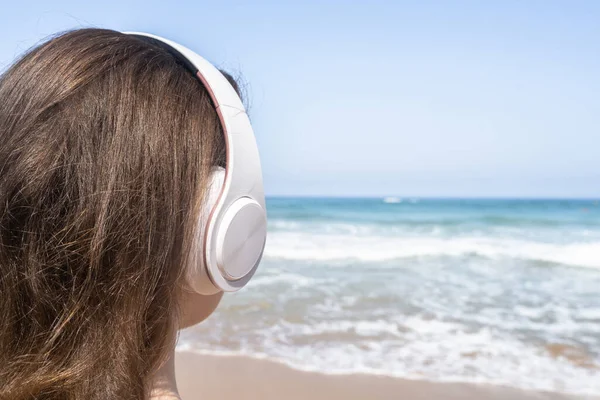 Alone woman on a beach in headphones listen music looking on the sea. Female relaxation at summer vacation. Back view