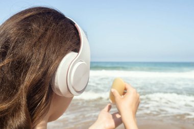 Kulaklıklı kadın, plajda denize bakan tenine güneş kremi sürerken müzik dinliyor. Yaz tatilinde kadın dinlenmesi. Arka plan