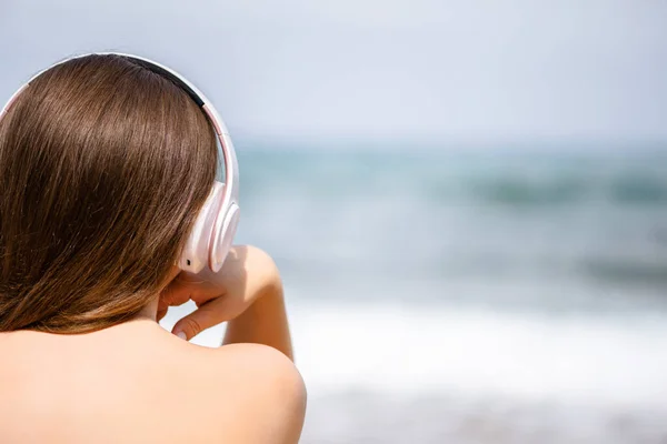 Back view of alone woman sitting on a beach in headphones listen music dreaming and looking on the sea. Female relaxation at summer vacation.