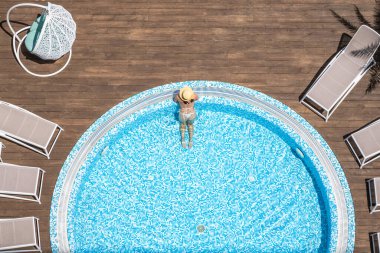 Yalnız, bikinili ve şapkalı güzel bayan jakuzide, tahta terasta yüzüyor. Yukarıdan insansız hava aracı görüntüsü. Tropik yaz lüks oteli