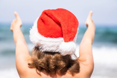 Noel deniz tatili. Noel Baba şapkalı mutlu kadının Cennet Sahili Adası kaçamağında baş parmağını göstermesi ve rahatlaması. Yeni Yıl