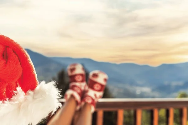 Woman in socks, boots and Christmas red hat sitting on a balcony in the mountains putting her feet on the wooden terrace railing