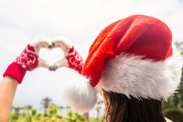 クリスマス海の休日 サンタクラスの帽子と手袋の幸せな女性のバックビュー手からハートの形を示し ヤシの木とパラダイスビーチの島でリラックス — ストック写真