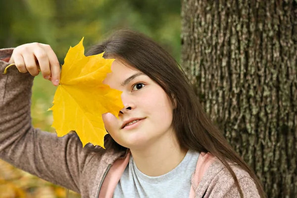 Portrait. The girl covers her face with a maple leaf. Young lady with a yellow leaf on a tree background. — Stock Photo, Image