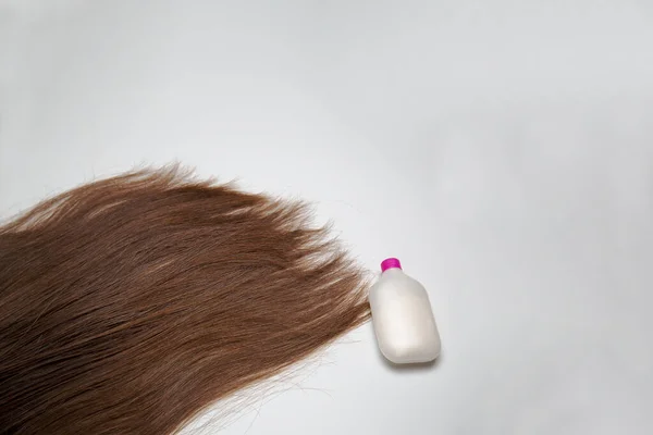 Thick, long, natural hair lies on a white surface. Brown hair on a white background. Top view. A white tube of shampoos, a bottle of balsam. Hair care and treatment.