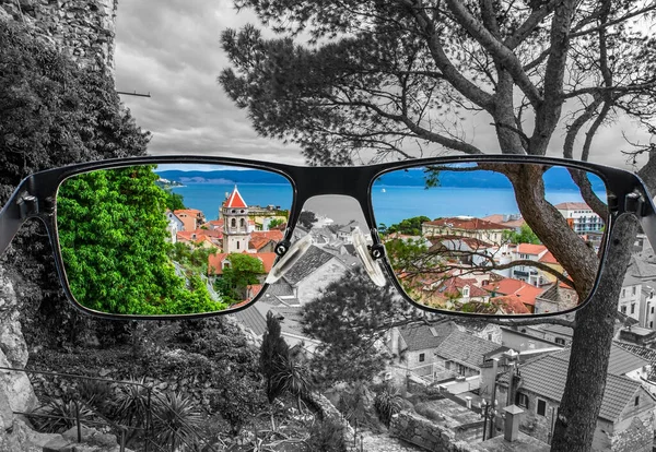 stock image Looking through glasses to colorful view of pine tree, old city center with red rooftops and blue see surrounded by black and white background. Different world perception. Optimism, hopefulness.