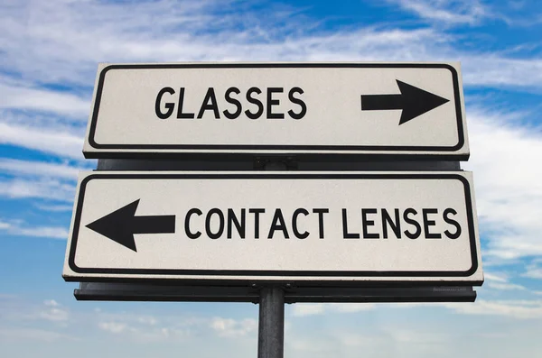 Glasses versus contact lenses road sign with two arrows on blue sky background. White two street sign with arrows on metal pole. Two way road sign with text.