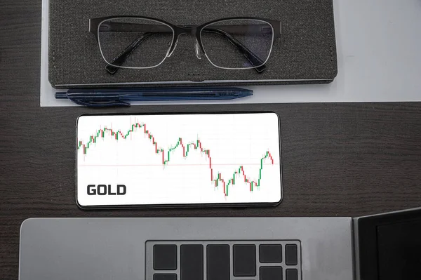 Buy or sell gold concept. Top view of stocks price candlestick chart in phone on table near laptop, notepad and glasses with inscription gold. Business, finance concept
