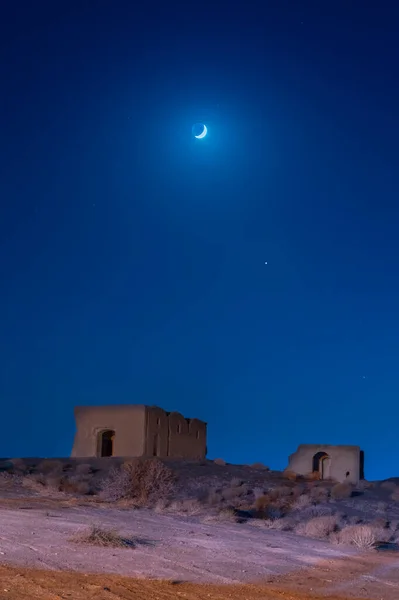 night view from ruins of two houses on a hill in desert with moon and stars under blue sky in iran