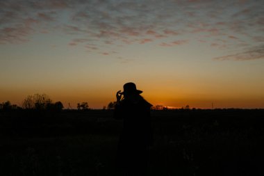 Siyah paltolu ve şapkalı genç şehvetli kadın akşam vakti altın sonbahar tarlasında duruyor..