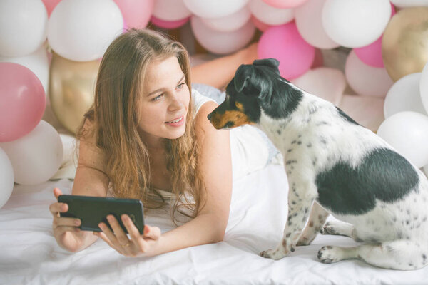 Lovely lady in pajama making selfie in her bedroom using phone and hug her dog. Indoor portrait  girl with baloons in morning. Husband surprising his wife.