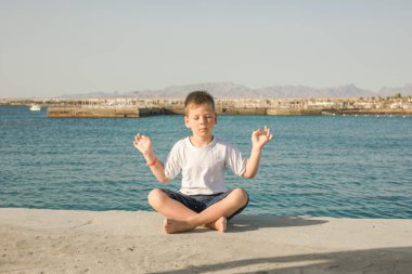 Sevimli, mutlu, gülümseyen küçük bir çocuk. Gözleri kapalı, açık denizde yoga ve meditasyon yapıyor. Yeni başlayan meditasyon, bir çocuğun konsantrasyon konseptini geliştirmek