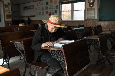 Amiş çocuk eski ahşap bir okul masasında elinde eski sınıf bir kitapla oturuyor..