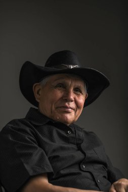 Siyah şapka ve gömlek giyen yaşlı bir adamın yakın plan, sade bir portresi. Nazik ve düşünceli bir ifadeyle hafifçe yukarı bakıyor. Yumuşak ışık yüzündeki kırışıklıkları vurguluyor, karamsar ve iç gözlemci bir atmosfer yaratıyor..