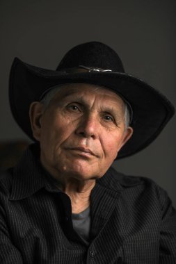 Siyah şapka ve gömlek giyen yaşlı bir adamın yakın plan, sade bir portresi. Nazik ve düşünceli bir ifadeyle hafifçe yukarı bakıyor. Yumuşak ışık yüzündeki kırışıklıkları vurguluyor, karamsar ve iç gözlemci bir atmosfer yaratıyor..