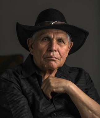 Siyah şapka ve gömlek giyen yaşlı bir adamın yakın plan, sade bir portresi. Nazik ve düşünceli bir ifadeyle hafifçe yukarı bakıyor. Yumuşak ışık yüzündeki kırışıklıkları vurguluyor, karamsar ve iç gözlemci bir atmosfer yaratıyor..