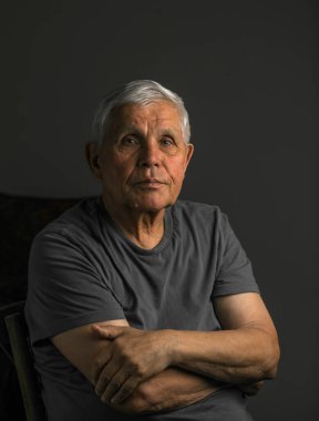 Siyah şapka ve gömlek giyen yaşlı bir adamın yakın plan, sade bir portresi. Nazik ve düşünceli bir ifadeyle hafifçe yukarı bakıyor. Yumuşak ışık yüzündeki kırışıklıkları vurguluyor, karamsar ve iç gözlemci bir atmosfer yaratıyor..