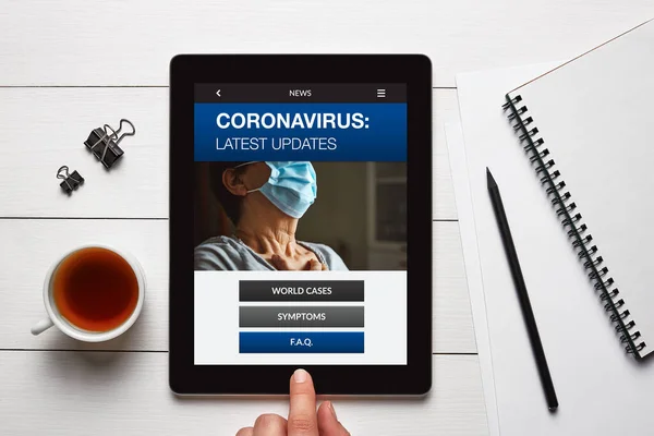 Coronavirus latest news concept on tablet screen with office objects on white wooden table. Top view