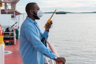 Güverte subayı ya da Birinci Subay geminin güvertesinde ya da koruyucu maskeli bir gemide. Elinde bir VHF telsiz telsizi var. Tekne iletişimi