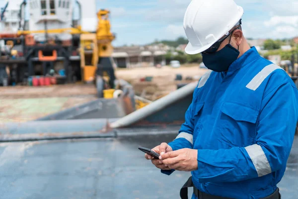 Deck officer with protective mask on the deck of a seagoing vessel, with cell phone