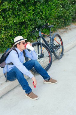 Bisikletinin yanında kahve içen yaşlı bir adam resmi.