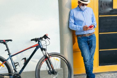 Koruyucu maskeli bir iş adamı bisikletinin yanındaki cep telefonunu kontrol ediyor.