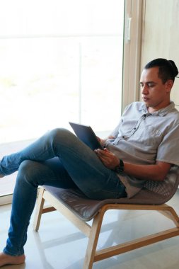 Evinin oturma odasında oturan bir adam, evden çalışmak için tablet kullanıyor.