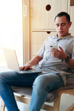 Oturma odasında otururken cep telefonu ve laptop kullanan bir adam.