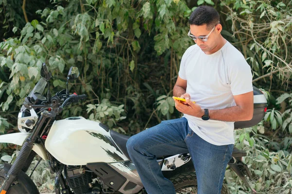 Young Hispanic man in white shirt and jeans checking cell phone near motorcycle outdoors