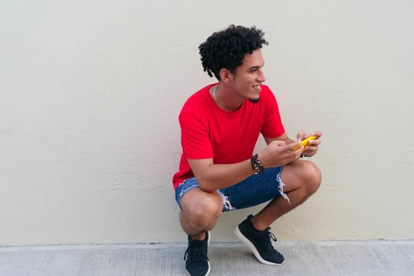 Jovem Rindo Agachado Frente Uma Parede Cinza Com Telefone Celular — Fotografia de Stock