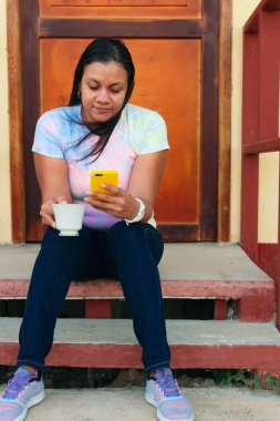 Evinin önünde oturan, kahve içen ve cep telefonunu kontrol eden güzel Latin bir kadın.