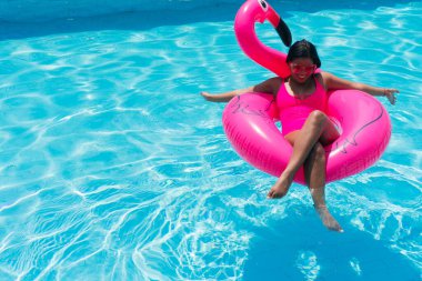 Smiling young woman in bikini relaxes on inflatable pink flamingo in swimming pool. Attractive woman in swimsuit lies in the sun on tropical vacation. Woman sunbathing at the resort.