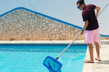 Man cleaning swimming pool of fallen leaves with net in summer