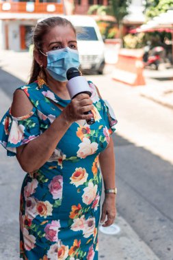 Tıbbi maskeli ve mikrofonlu olgun bir kadın gazeteci şehir sokaklarında çalışıyor. Antivirüs koruması