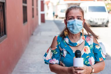 Tıbbi maskeli ve mikrofonlu olgun bir kadın gazeteci şehir sokaklarında çalışıyor. Antivirüs koruması