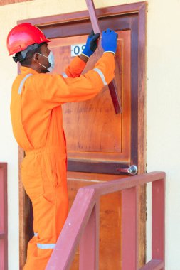 Tulumlu bir işçi, kapı ölçüleri alıyor. Koruyucu maskeli bir adam kapıyı tamir ediyor..