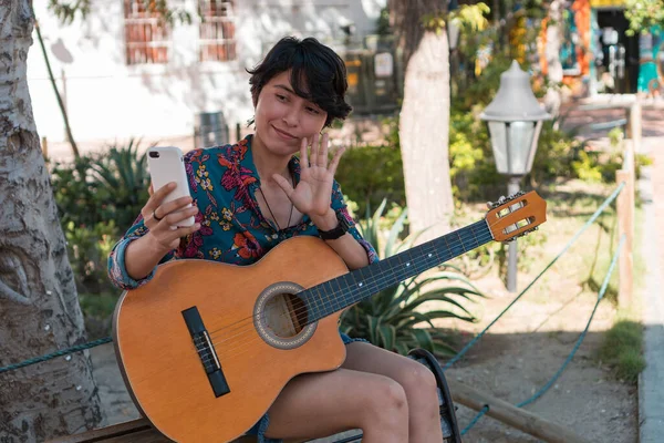 Woman with guitar making a video call while sitting in a park