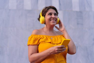 Sokakta kulaklıkla müzik dinlerken cep telefonu kullanan olgun Latin bir kadın.