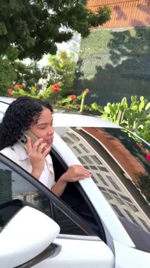 Kıvırcık saçlı genç bir kadın beyaz bir arabada akıllı telefonuyla hoş bir sohbet ediyor.