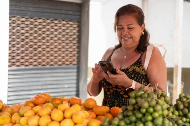 Açık hava meyve pazarında akıllı telefonunu kullanan bir kadın gülümsüyor.