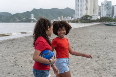 Sahilde yürüyen iki mutlu kız, ellerinde bir futbol topu, arka planda şehir