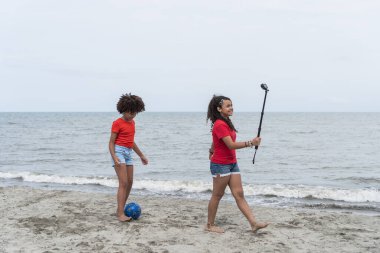 Genç kızlar plaj tatilinden hoşlanıyor, biri futbol oynuyor, diğeri de selfie çubuğuyla kayıt yapıyor.