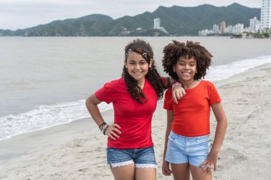 Arka planda şehir manzarası olan bir plajda dururken gülümseyen genç kızlar.