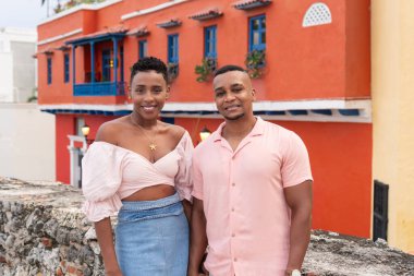 Eski bir şehirde renkli binaların önünde dikilen siyah çift gülümsüyor.