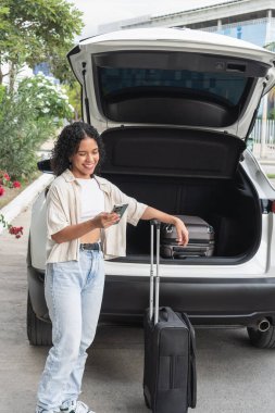 Akıllı telefon kullanarak gülümseyen genç bir kadın. Bagajın yanında duruyor.