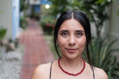 Kameraya bakan genç bir kadın, bir burun halkası ve dışarıda kırmızı bir kolye takıyor.