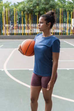 Elinde basketbol topu olan genç bir kadın, açık hava sahasına odaklanmış.