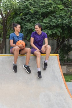 İki gülümseyen kadın arkadaş kaykay parkında oturmuş, konuşuyorlardı ve ellerinde basketbol topu vardı.